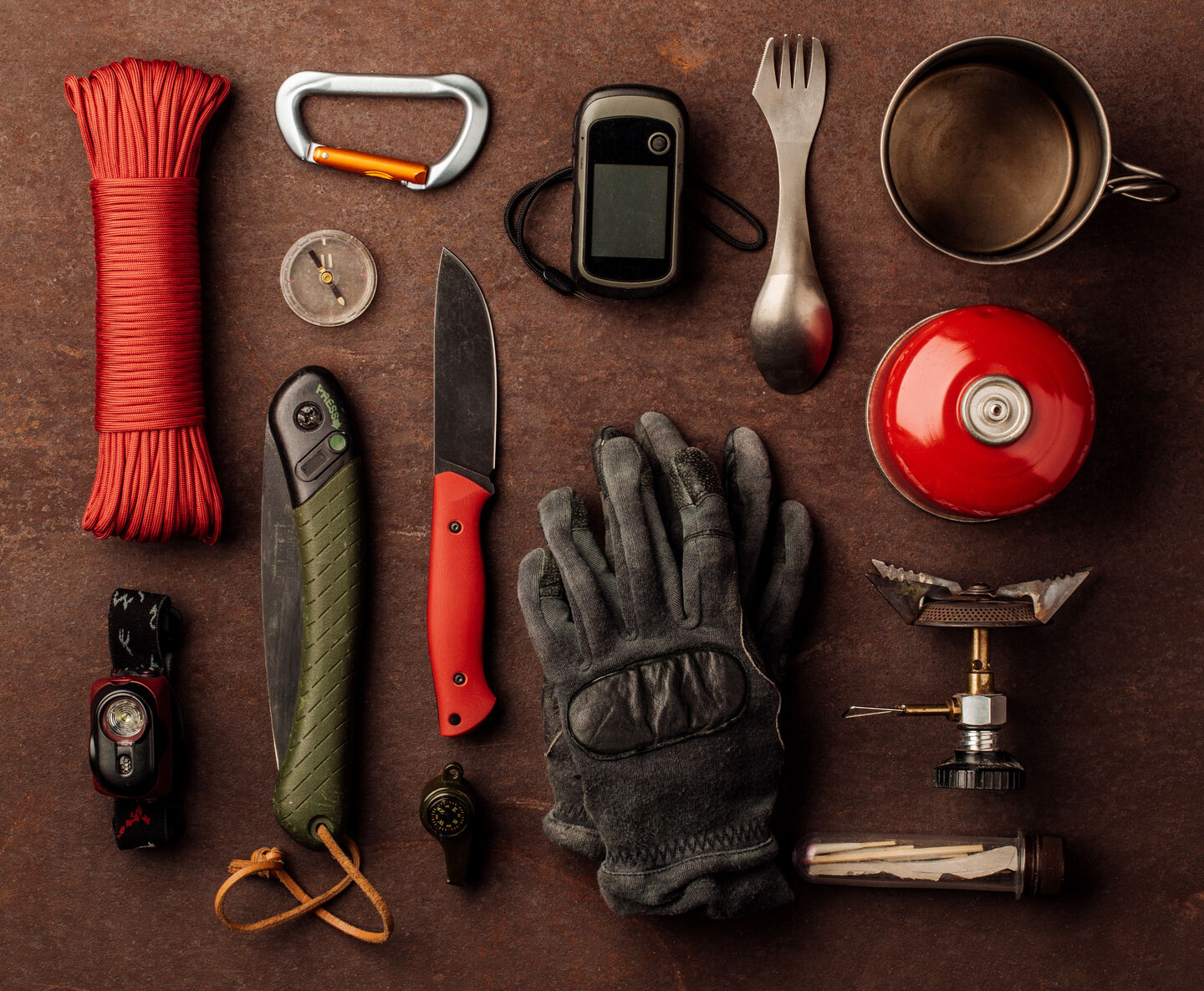Two hand knives, a wrapped cord, a pair of gloves, a compass, and camping stove components lie on a brown surface.